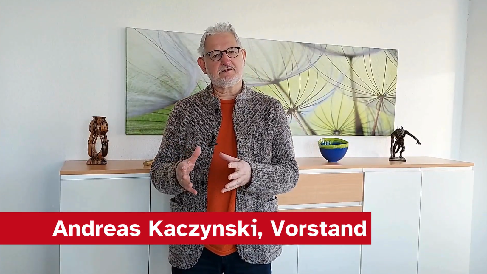 Ein Mann namens Andreas Kaczynski steht in einem hellen Raum vor einem Sideboard. Er trägt eine graue Jacke und ein oranges T-Shirt. Im Hintergrund ist ein großes Dekobild mit Pflanzenmotiven sowie einige dekorative Objekte auf dem Sideboard zu sehen.