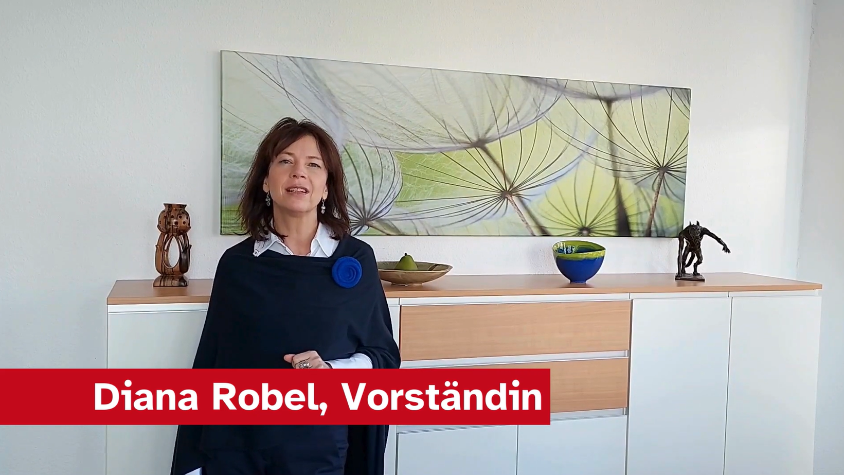 Eine Frau steht vor einem modernen Sideboard, auf dem Dekorationsobjekte und eine Schüssel stehen. Im Hintergrund ist ein großes, farbenfrohes Bild von Pflanzen zu sehen. Sie trägt einen eleganten schwarzen Umhang und lächelt. Am unteren Rand steht der Text: „Diana Robel, Vorstandin“.
