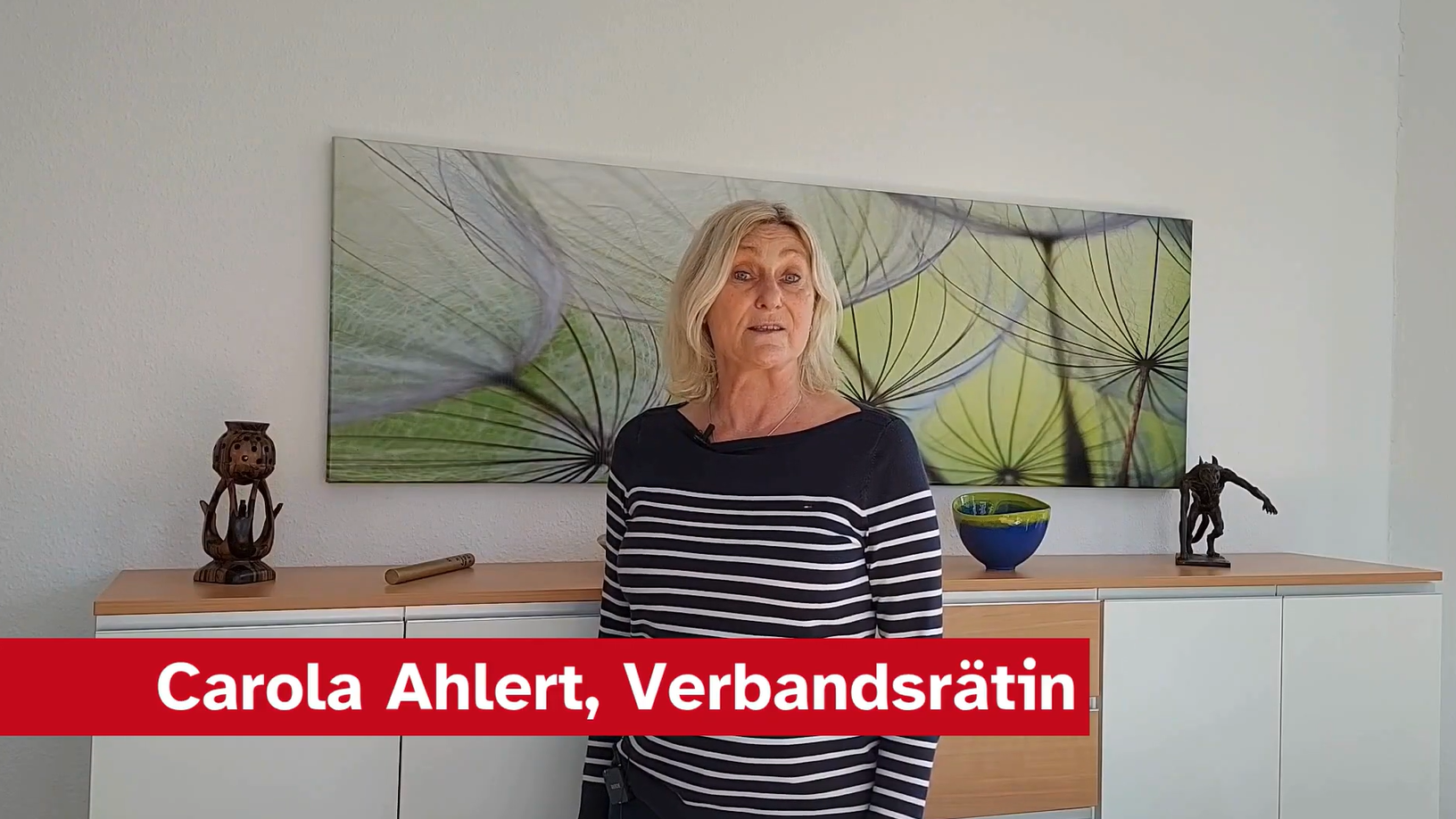 Eine Frau mit langen, blonden Haaren steht vor einer hellen Wand. Sie trägt ein gestreiftes Oberteil und spricht in die Kamera, während im Hintergrund ein modernes Gemälde sowie einige Dekorationsobjekte auf einem Sideboard sichtbar sind.
